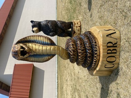 A magnificent chainsaw carving of a cobra, its hood flared and mouth open, sits atop a circular base carved with the word "COBRAS." The cobra is painted in shades of gold, brown, and blue. To the right and slightly behind the cobra is another ch